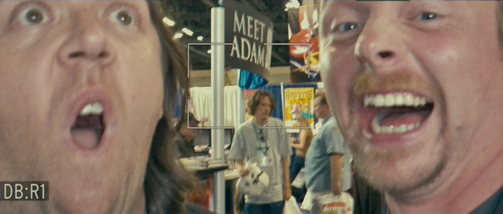 Paul (2011) movie still: closeup — A bustling convention center hall is filled with booths and attendees in the distance. Two men take a close-up…