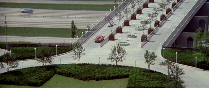 Brewster McCloud (1970) movie still: wide — A wide exterior shot depicts a concrete pedestrian ramp leading to a large building. An orange car…