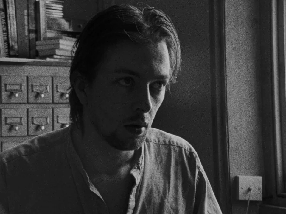 Following (1998) movie still: closeup — A man sits in front of a wooden card catalog cabinet inside a library. Books are stacked on top of the…