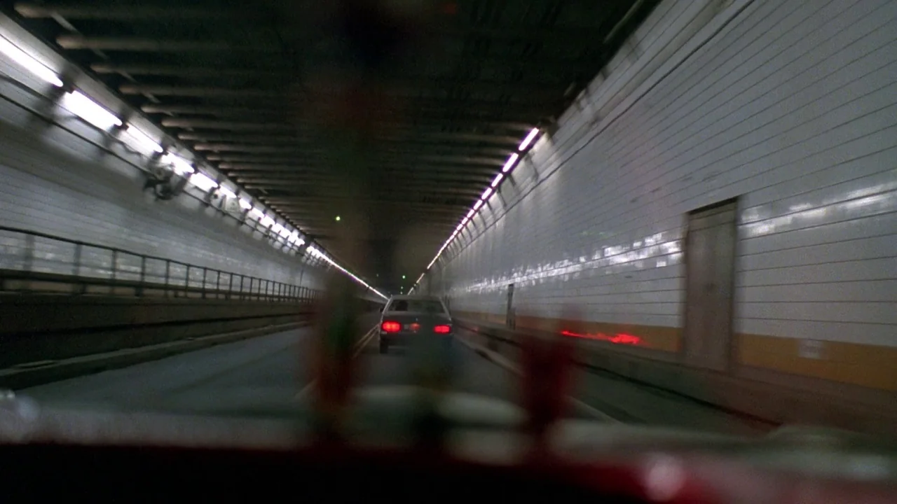 Something Wild (1986) movie still: wide — A long, white-tiled vehicular tunnel recedes into the distance under rows of fluorescent lights. A dark…