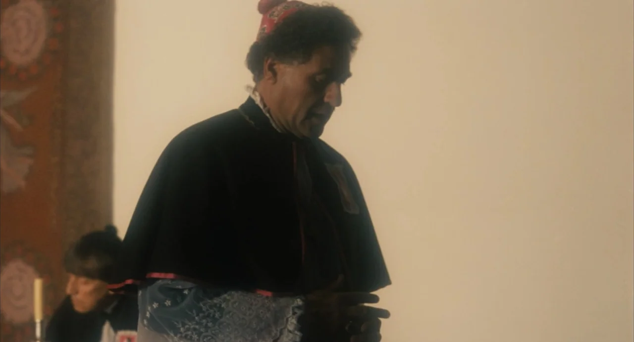 Camila (1984) movie still: medium — An interior space with a plain, off-white wall and a decorative fabric tapestry hanging. A man in formal…