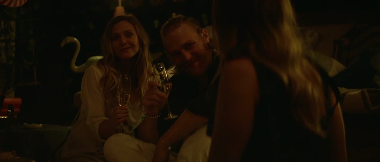 Ingrid Goes West (2017) movie still: medium — A dimly lit indoor space features a man and woman holding champagne flutes together. The background is…