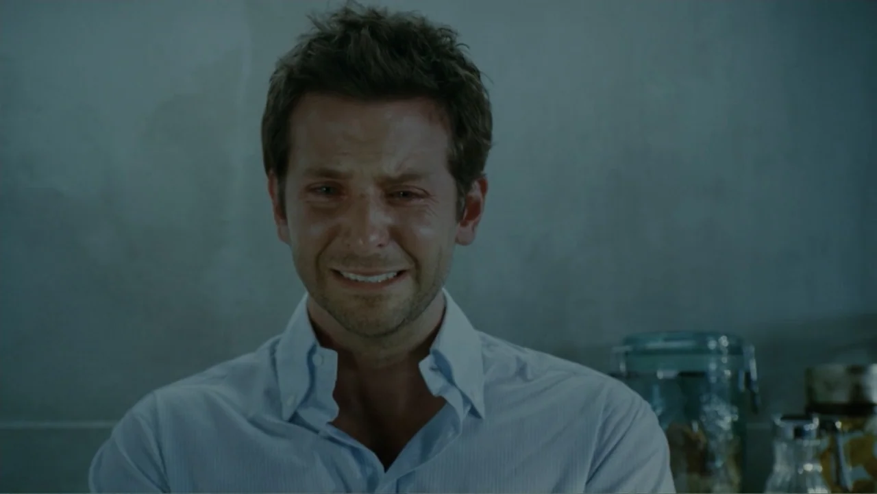 The Words (2012) movie still: closeup — An interior space features a gray tiled wall and blurred kitchen storage jars. A man in a blue dress shirt…