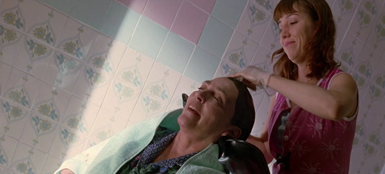 Volver (2006) movie still: medium — A residential bathroom features wall-to-wall ceramic tiles with a light floral pattern. A woman washes the hair…