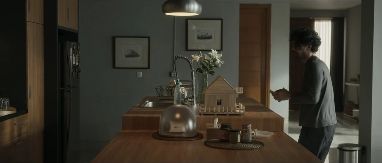 Innocent Vengeance (2023) movie still: medium — A modern kitchen island made of wood sits beneath a circular pendant light fixture. A man in a…