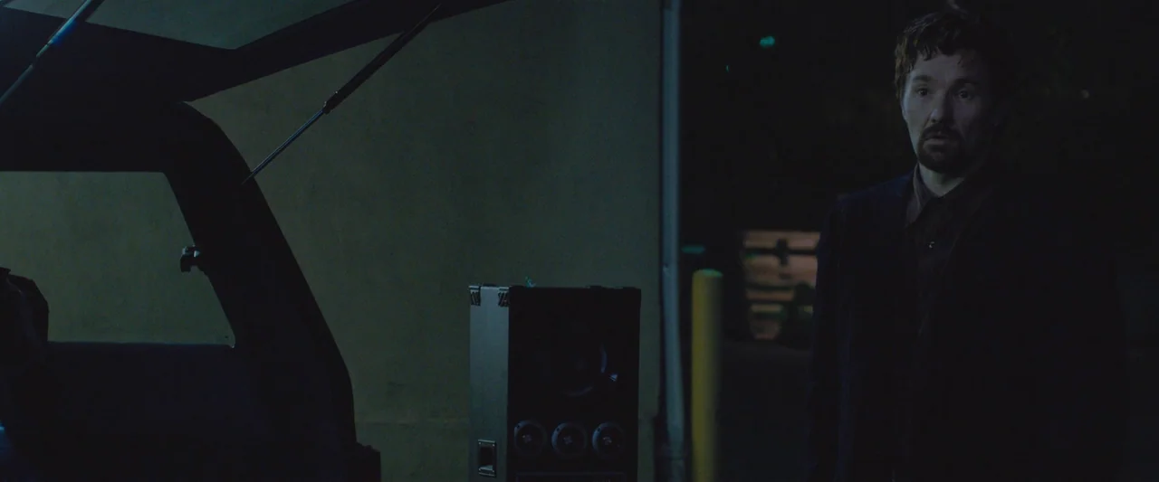 The Gift (2015) movie still: medium — A dark, sparsely lit parking lot with a car trunk open in the foreground. A man wearing a dark jacket stands…