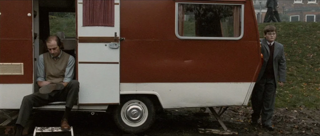 Tinker Tailor Soldier Spy (2011) movie still: wide — A parked vintage two-tone red and white camper trailer sits on a muddy slope. An adult male…