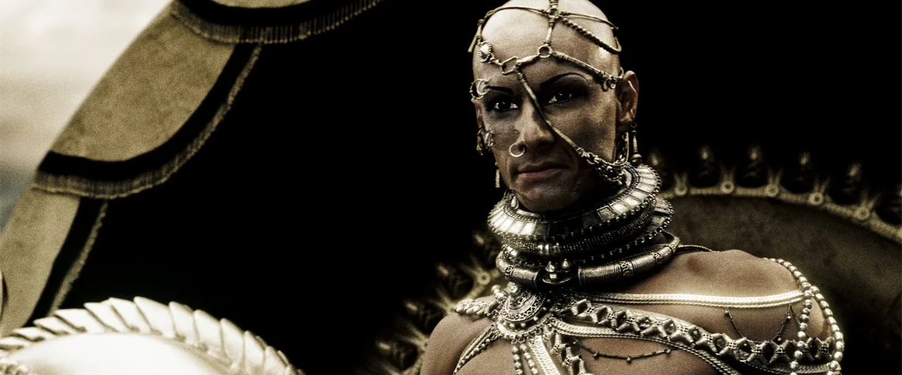 300 (2007) movie still: medium — A high-fantasy royal chamber features dark architectural elements behind a seated figure. A man in heavy golden…
