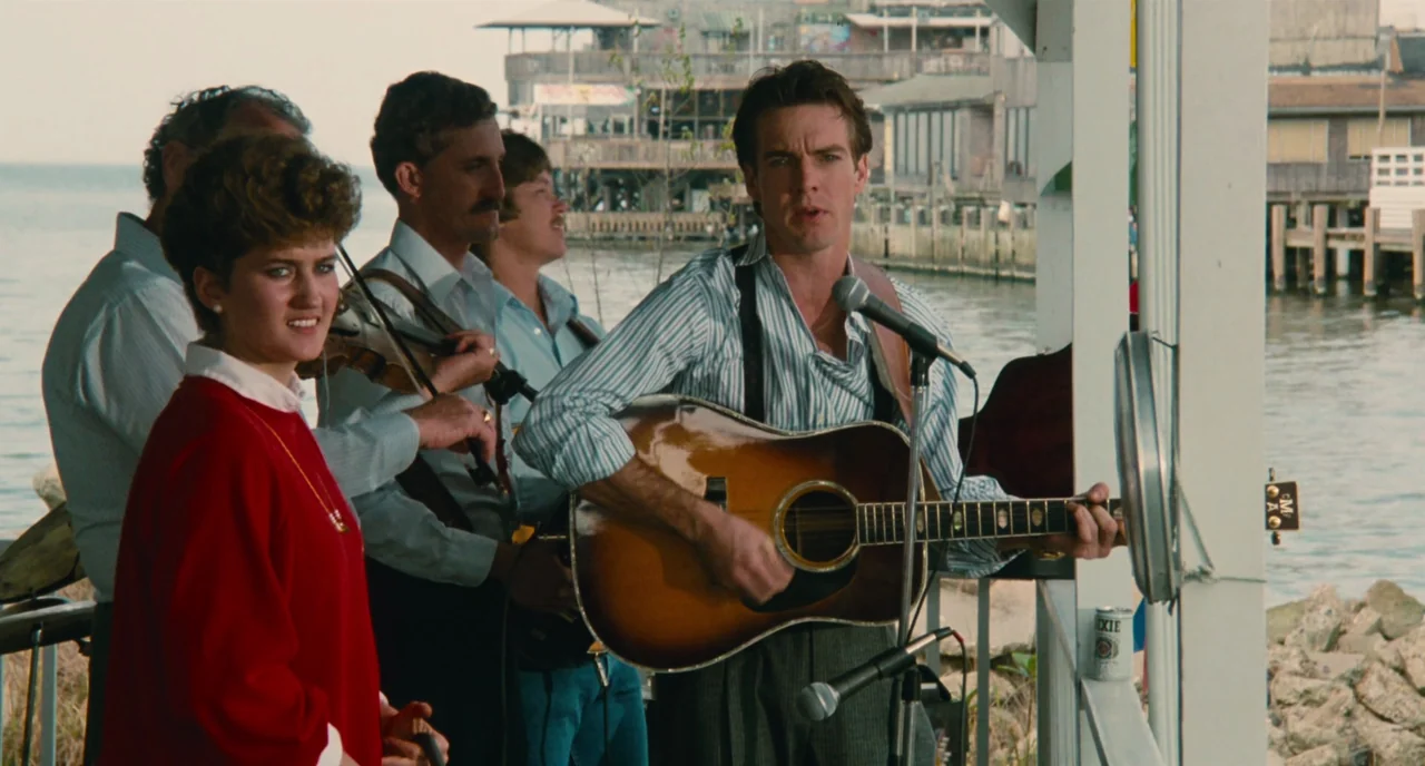 The Big Easy (1986) movie still: medium — A musical group performs on a wooden dock overlooking a calm, grey coastal waterway. The band includes a…
