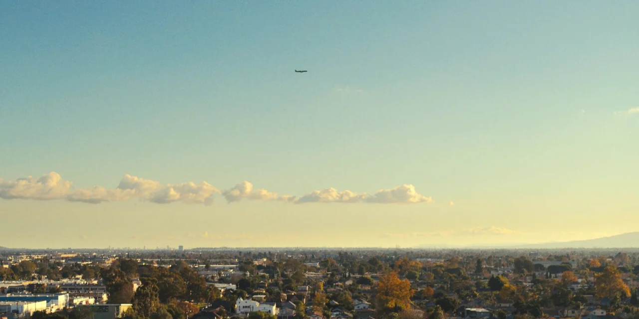 Silent Night (2023) movie still: establishing — An aerial view overlooks an expansive suburban city landscape under a vast blue sky. A distant small…