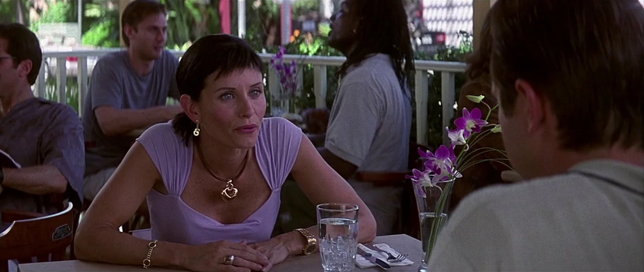 Scream 3 (2000) movie still: medium — An outdoor restaurant seating area with white wooden railings and partial foliage. A woman in a lilac top sits…