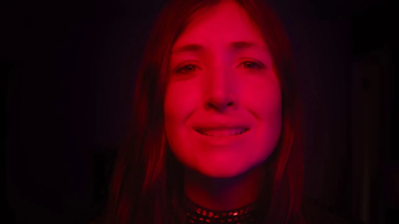She Dies Tomorrow (2020) movie still: closeup — A woman stands in a dark room illuminated by a strong red light. She looks directly at the camera…