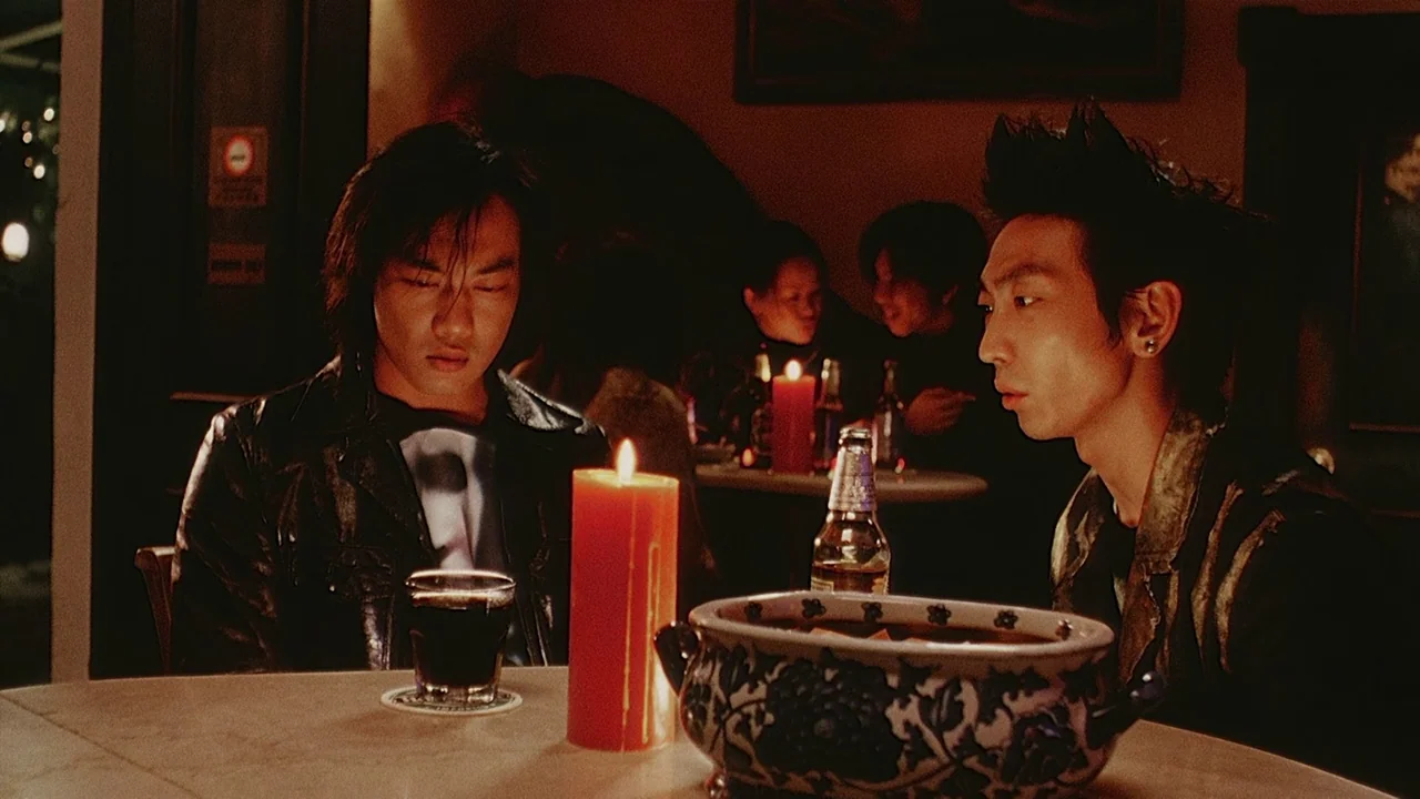 Visible Secret (2001) movie still: medium — A dimly lit restaurant interior features two men seated at a circular table. A candle, a drink glass,…