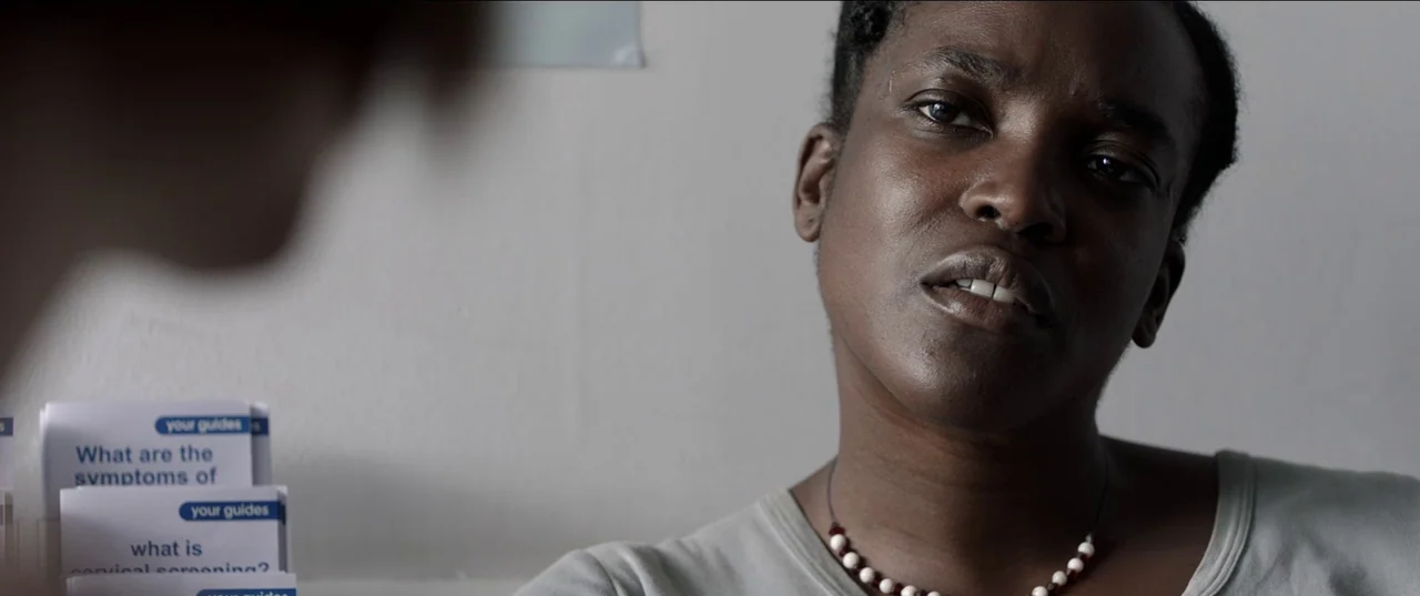 His House (2020) movie still: closeup — A medical office with a plain white wall and information brochures. A woman in a grey shirt listens to an…
