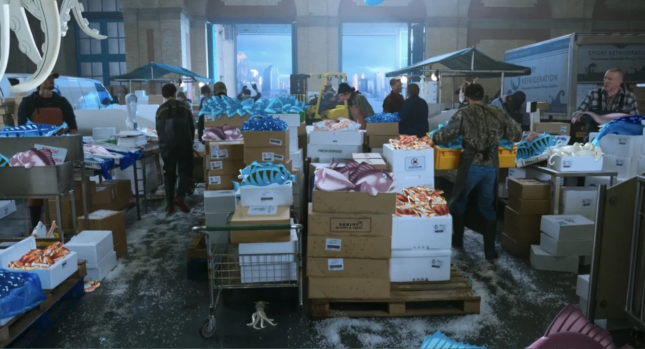 Tom & Jerry (2021) movie still: wide — A busy industrial fish market floor is filled with stacks of cardboard shipping boxes. Workers stand among…
