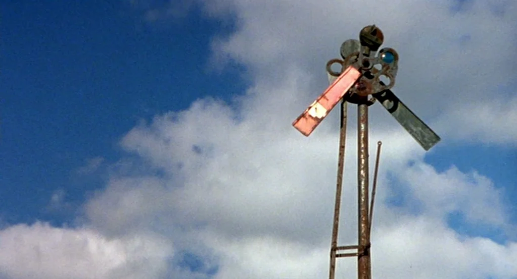 The Station Agent (2003) movie still: wide — A metal wind-pump stands against a bright blue sky filled with white clouds. The structure is made of…