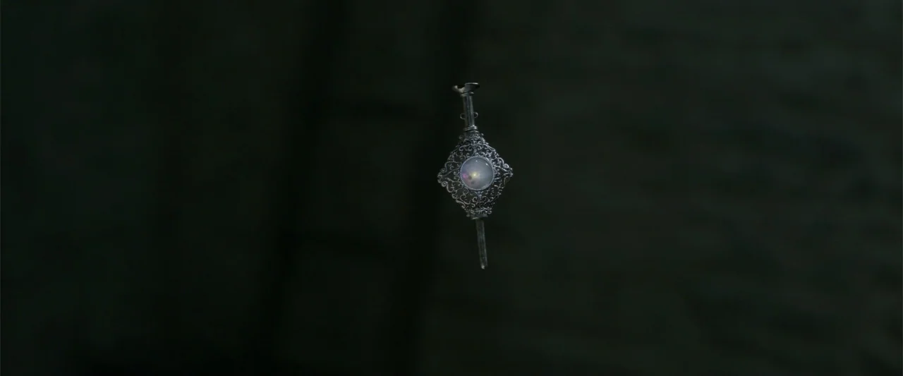Fantastic Beasts: The Crimes of Grindelwald (2018) movie still: closeup — A ornate silver pin hangs suspended against a pitch black background. The…