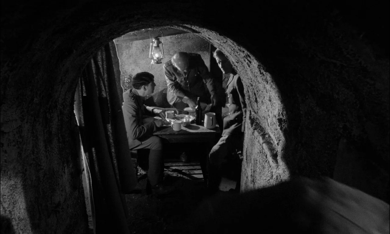 King & Country (1964) movie still: medium — A small underground space with rough hewn earth walls and an arched entryway. Three uniformed soldiers…