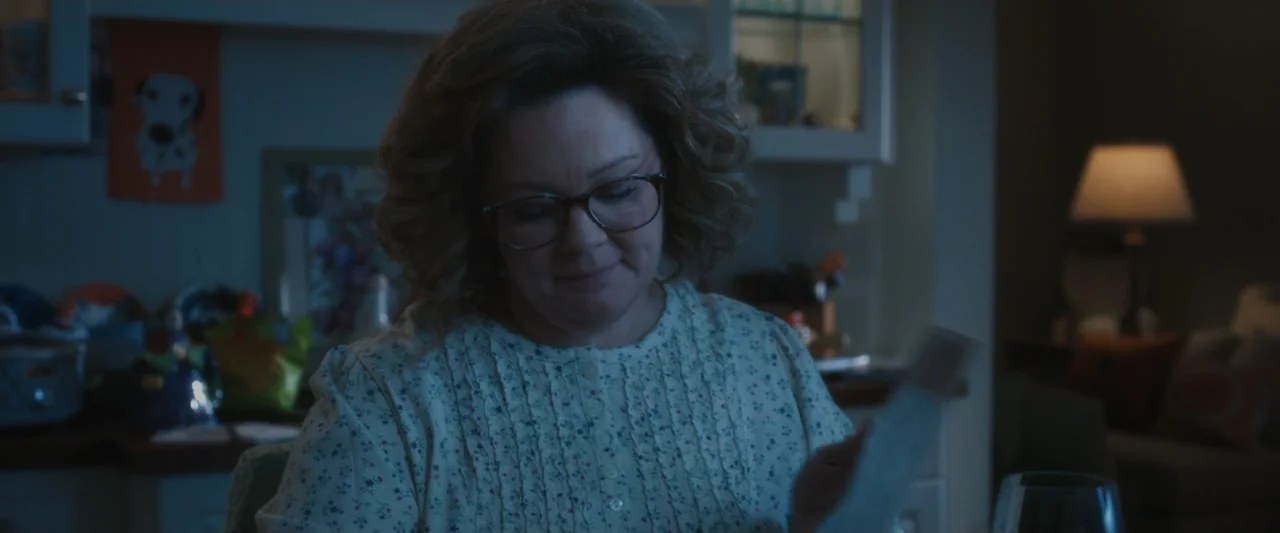 Life of the Party (2018) movie still: medium — A woman in a floral blouse sits in a darkened kitchen at night. She holds a small object in her hands…
