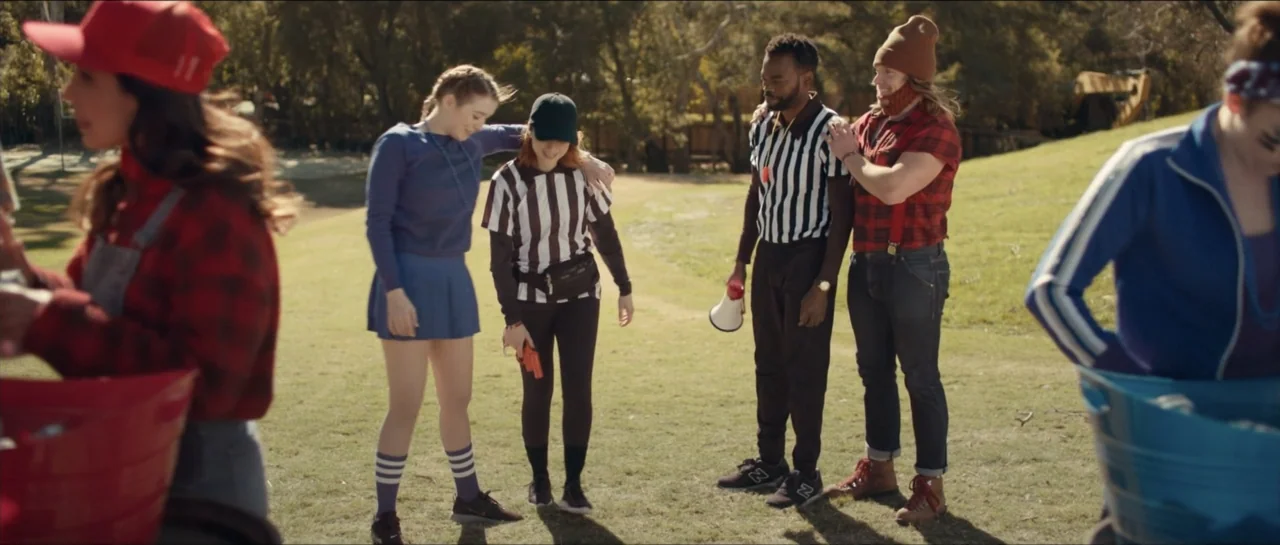 We Broke Up (2021) movie still: fullbody — A sunny grassy park area features several people in casual and referee costumes gathered. The group…
