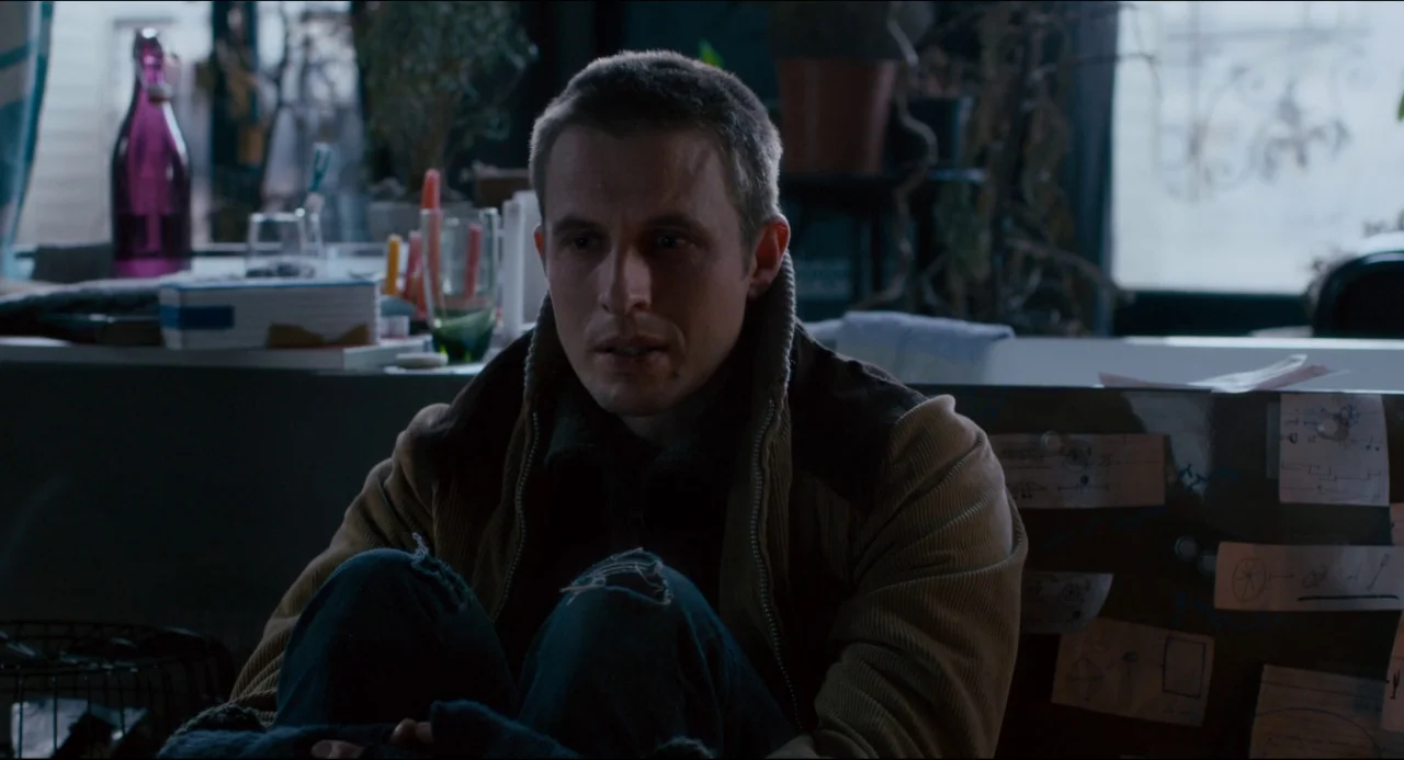 The Night Eats the World (2018) movie still: medium — A man sits on the floor of a cluttered apartment near a large window. He rests with his knees…
