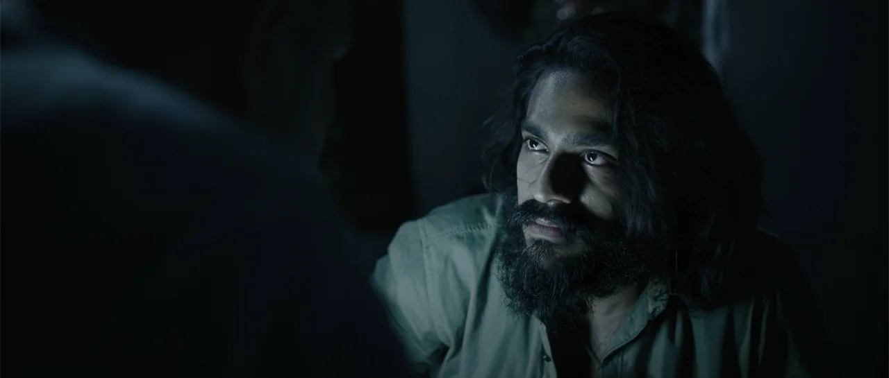 Nishaanchi 2 (2025) movie still: medium — A dark, minimally lit interior space with a person in the foreground. A man with dark hair and a beard…