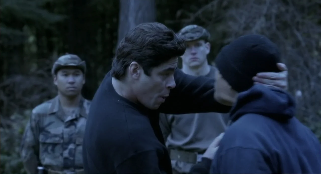 The Hunted (2003) movie still: medium — A group of men stand in a dense forest with tall trees and foliage. One man grips the back of another man's…