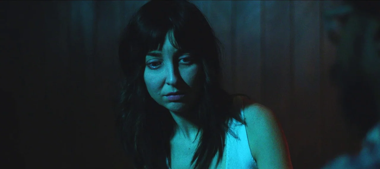 Fuzzy Head (2023) movie still: closeup — A dimly lit indoor space featuring a woman with dark hair in the foreground. She gazes downward while…
