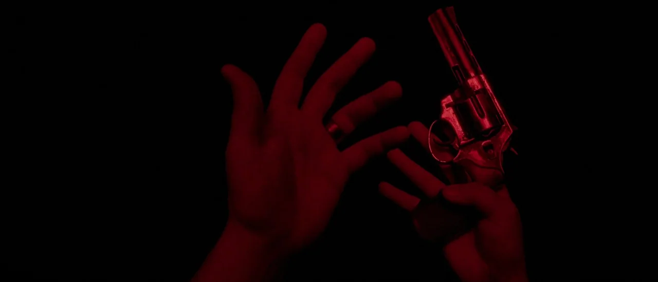 I Trapped The Devil (2019) movie still: closeup — A hand holding a metallic revolver is framed against a void of deep black. The scene is saturated…