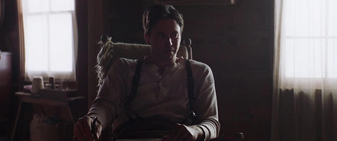 The Wind (2018) movie still: medium — A dimly lit room with wooden walls and two windows letting in daylight. A man sits in a rocking chair wearing…