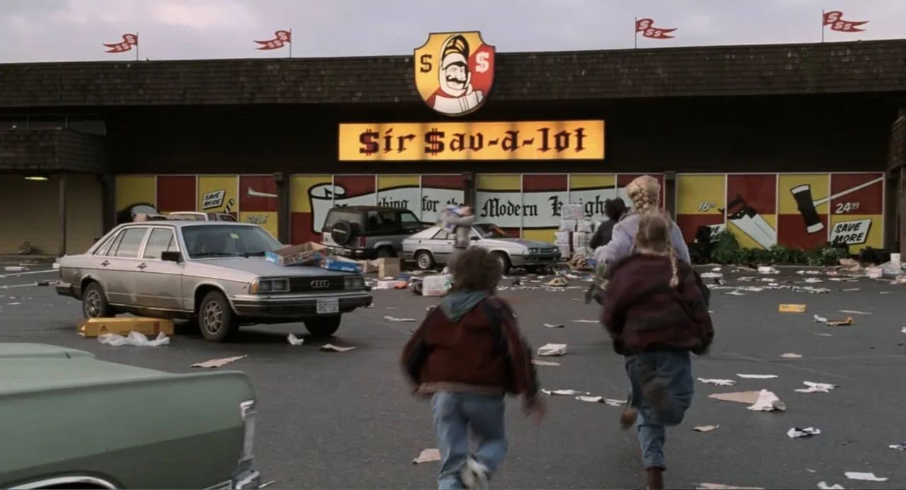 Jumanji (1995) movie still: wide — A cluttered asphalt parking lot sits before a retail storefront with large graphic signage. Two children run…