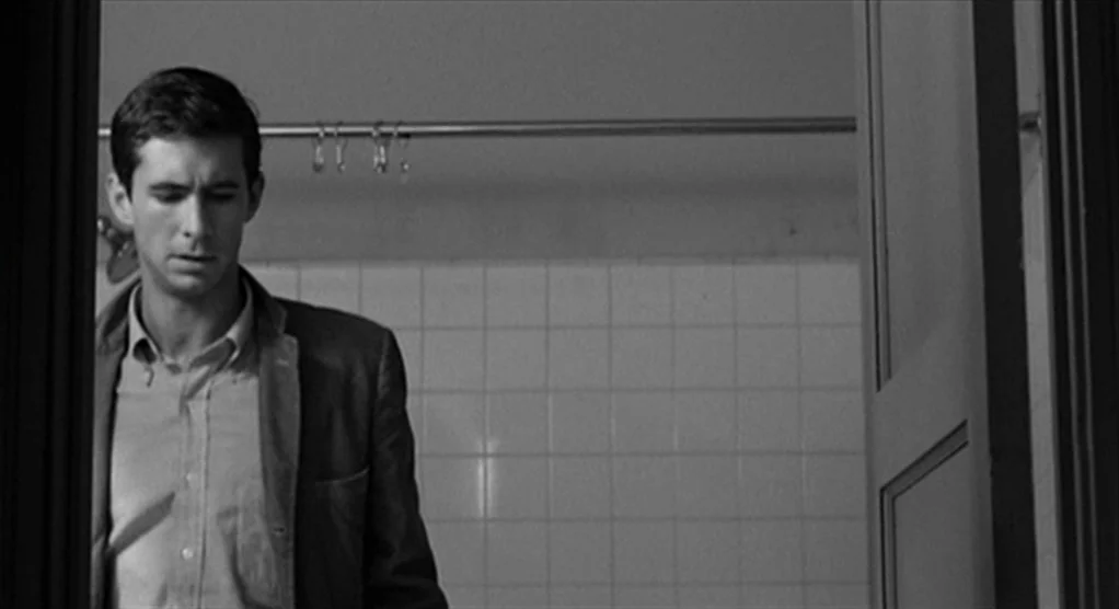 Psycho movie still: medium — A man stands in a tiled bathroom with a metal shower curtain rod behind him. He wears a button-down shirt and blazer…