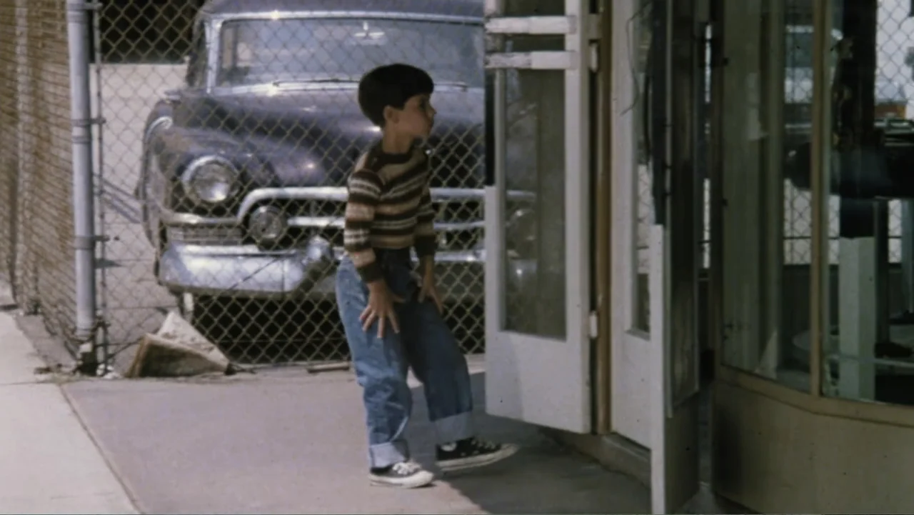 Aunt Mary (1979) movie still: fullbody — A sidewalk exterior features a chain-link fence, a parked vintage car, and a glass door. A young boy…