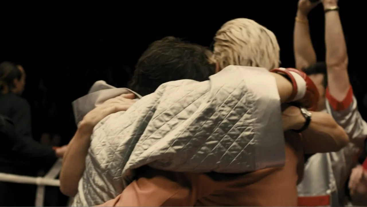 Bleed for This (2016) movie still: medium — Two individuals embrace inside a dimly lit professional athletic competition space. The two figures are…