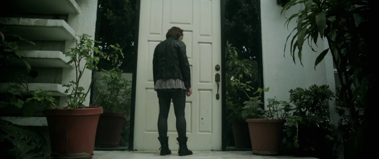Here Comes The Devil (2012) movie still: fullbody — A white front door is framed by white stucco walls and large terracotta pots. A woman stands in…
