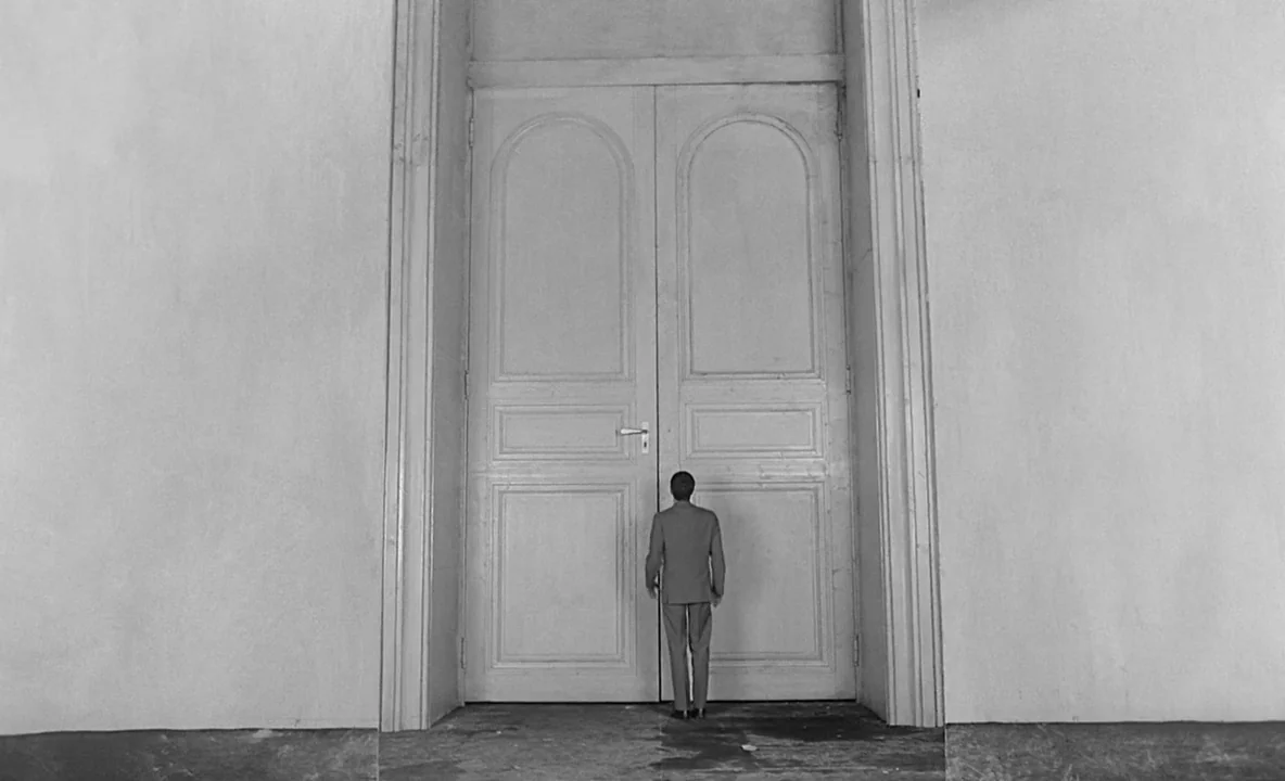 The Trial (1962) movie still: wide — A man stands centered before massive, floor-to-ceiling double doors within a minimalist stone corridor. He is…
