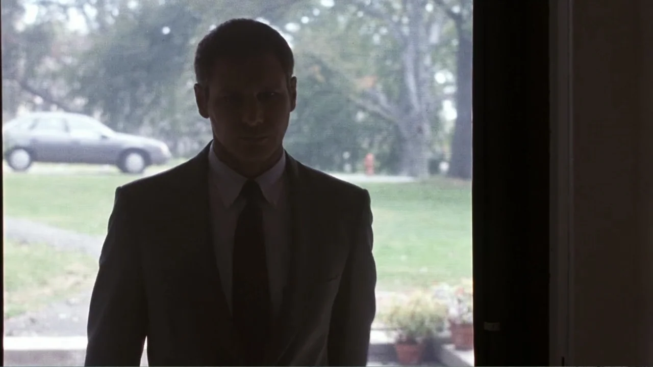 Presumed Innocent (1990) movie still: medium — A man in a dark suit stands in a residential doorway looking outward. The view behind him shows a…