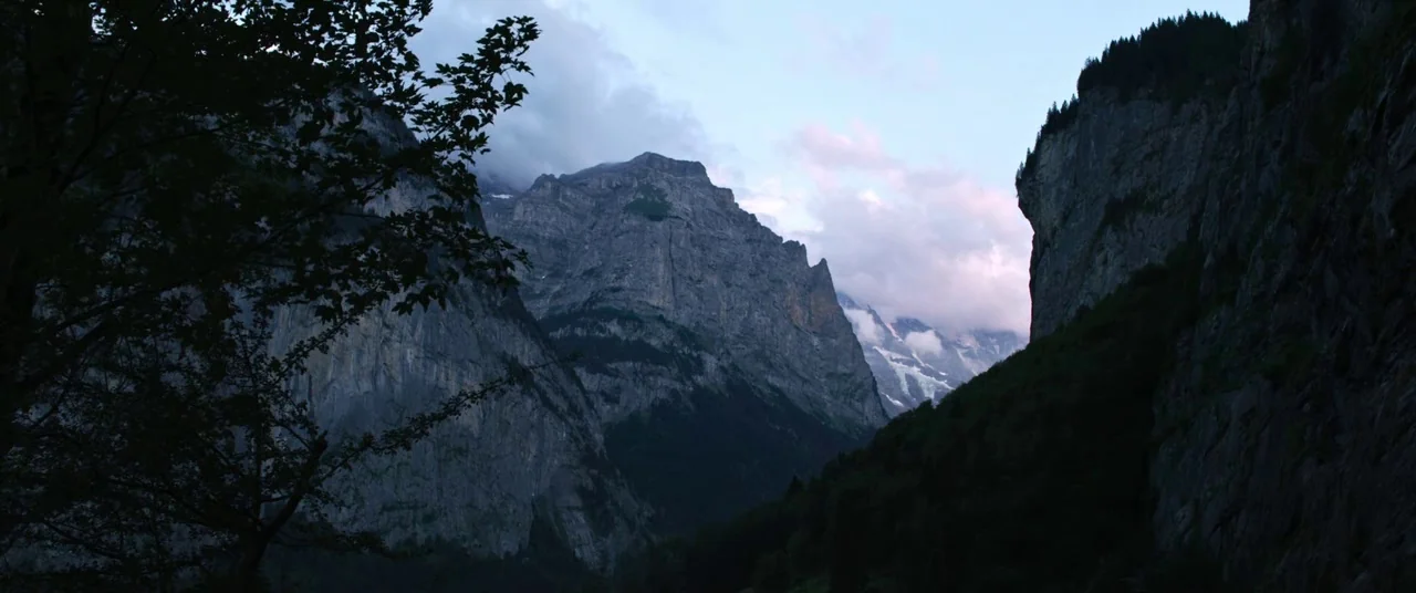 A Hidden Life (2019) movie still: establishing — A deep valley is flanked by massive, craggy rock faces and pine forests. Wispy clouds partially…
