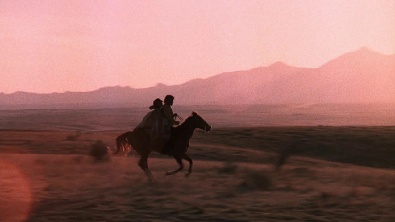 A Star Is Born (1976) movie still: wide — A vast, open desert landscape stretches out under a vibrant, hazy sunset sky. Two people ride a single…