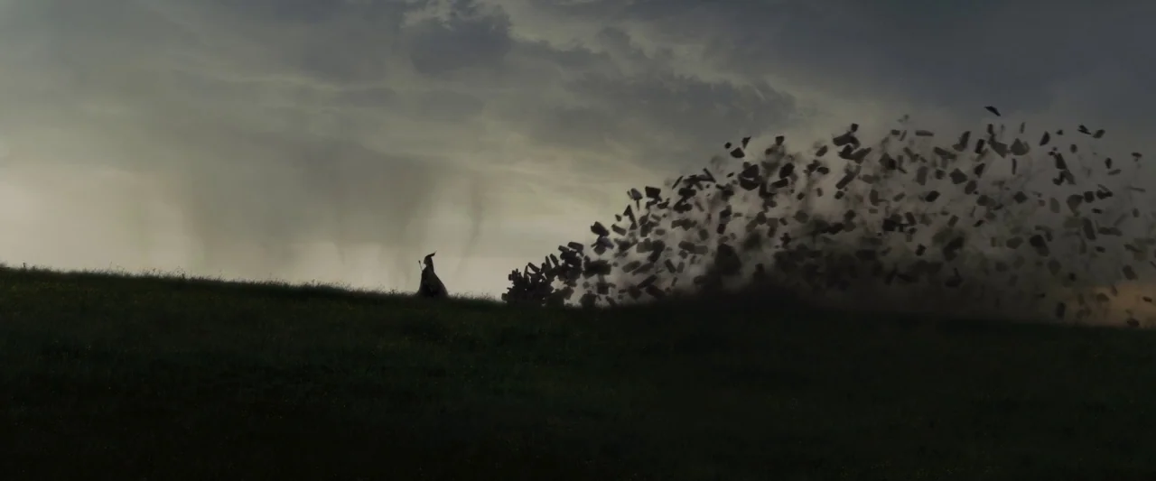 Maleficent (2014) movie still: wide — A dark, silhouetted figure stands atop a grassy hill under an overcast sky. A massive plume of dark debris and…