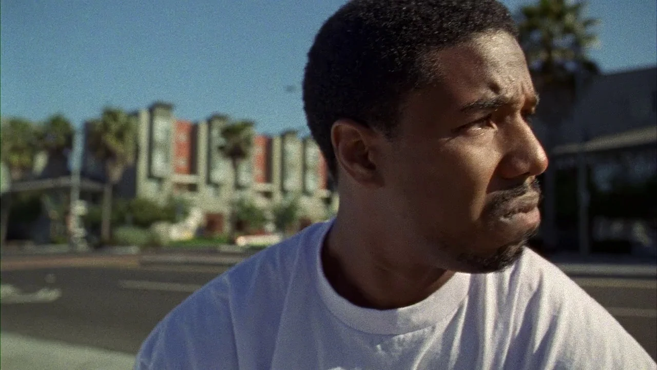 Fruitvale Station (2013) movie still: closeup — A man stands outdoors with modern low-rise buildings and palm trees in the background. He is…