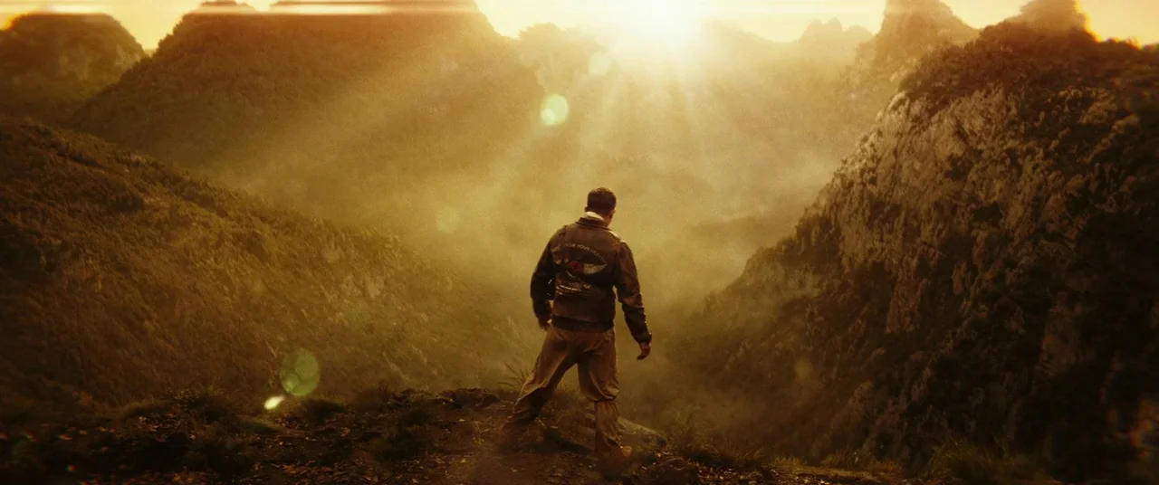 Kong: Skull Island (2017) movie still: wide — A rugged mountain range with steep slopes and hazy atmosphere under a bright sun. A man stands on the…