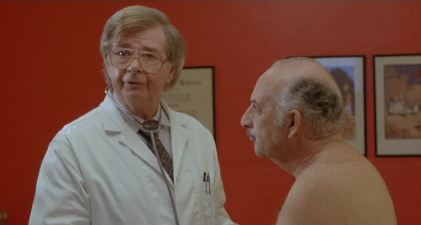 Scanners (1981) movie still: medium — A bright red room decorated with framed art hangs behind a doctor and patient. The doctor listens to the…