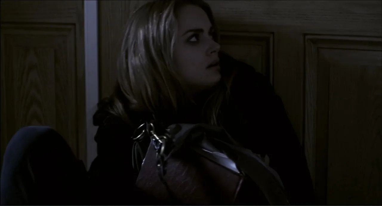 The Secret Village (2013) movie still: medium — A woman sits on the floor against a paneled wall in a dim space. She gazes to the side while holding…