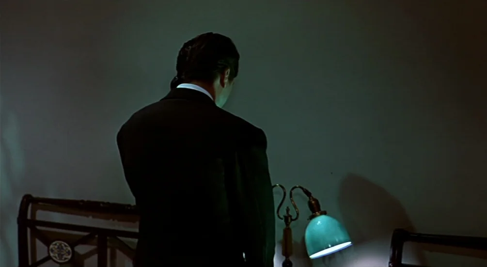 The Man Who Knew Too Much (1956) movie still: medium — A dark hotel room with minimal furniture and a plain wall. A man in a dark suit stands facing…