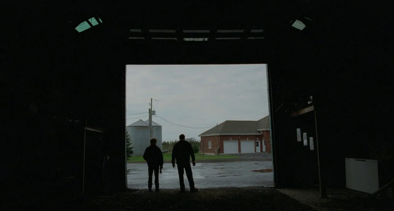 Tom at the Farm (2013) movie still: wide — The interior of a dark, cavernous barn looks out toward a rural house. Two men stand in the open doorway,…