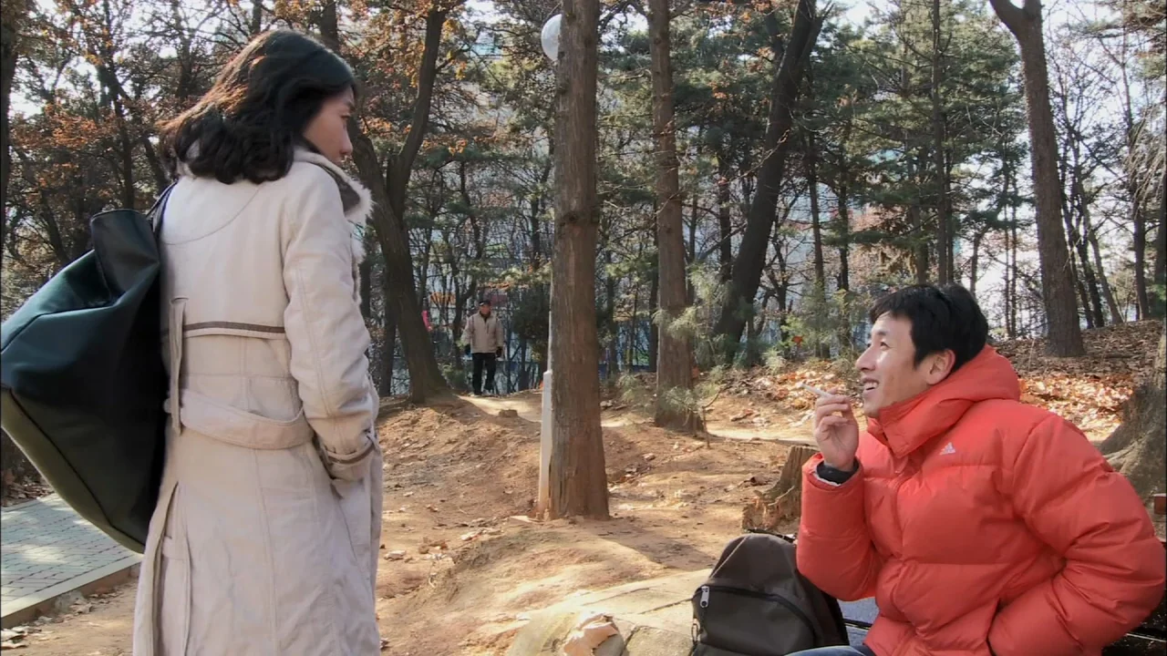 Oki’s Movie (2010) movie still: fullbody — A park setting with numerous tall, thin trees scattered across dirt pathways and dry leaves. A man in an…
