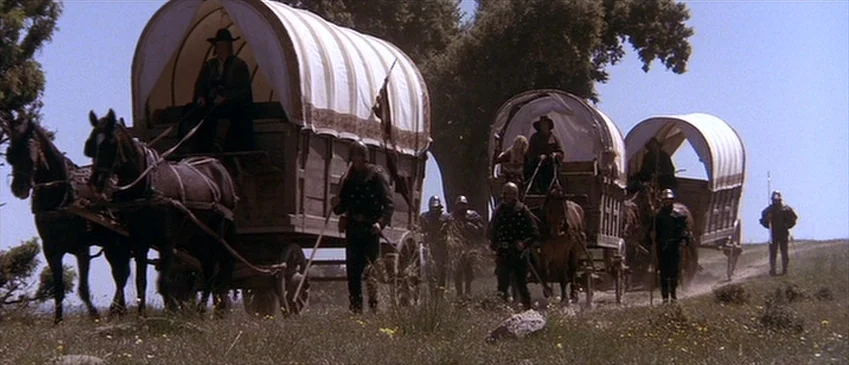 Flesh+Blood (1985) movie still: wide — Three horse-drawn covered wagons move across an open field on a dirt track. Several individuals dressed in…