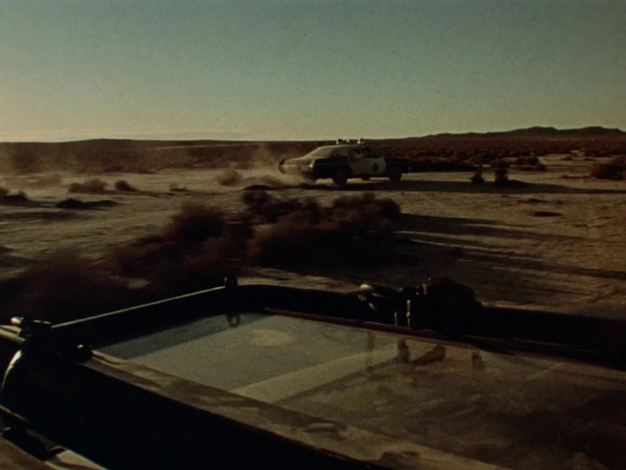Punishment Park (1971) movie still: wide — A flat, barren desert landscape stretches to the distant horizon under a clear sky. A police cruiser…