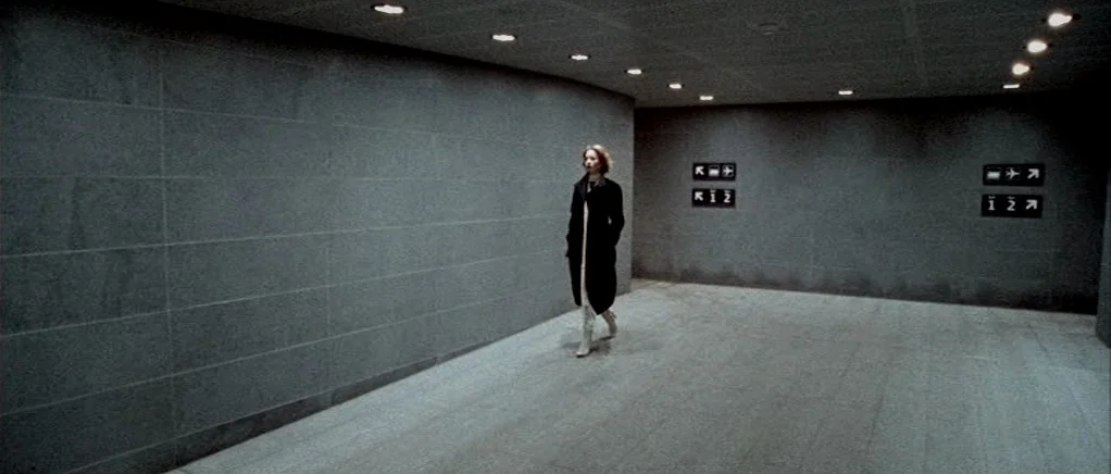Reconstruction (2003) movie still: wide — A dark, modern concrete terminal hallway features smooth grey walls and minimalist signage. A woman in a…